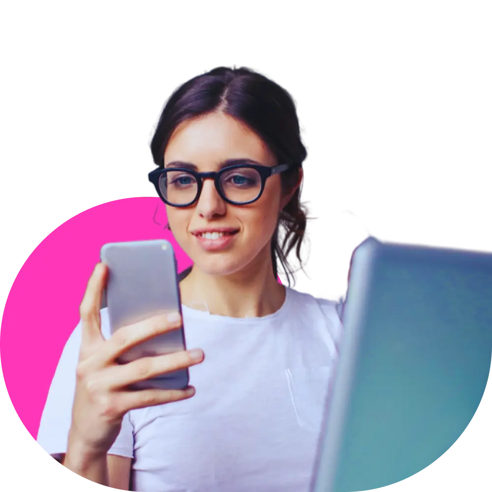 Woman wearing glasses, holding a smartphone and smiling while using a laptop