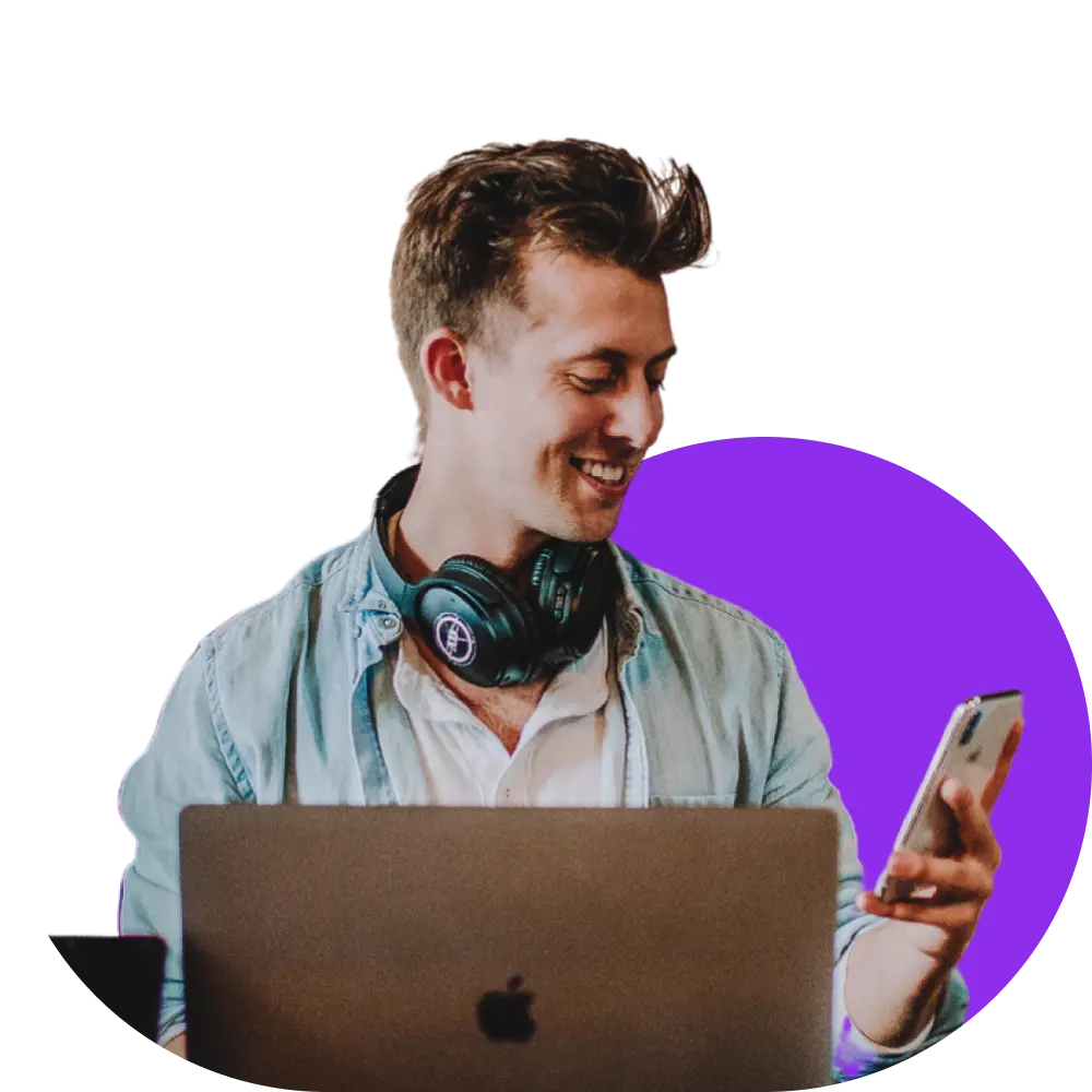 Man with headphones around his neck, smiling at his smartphone while working on a laptop